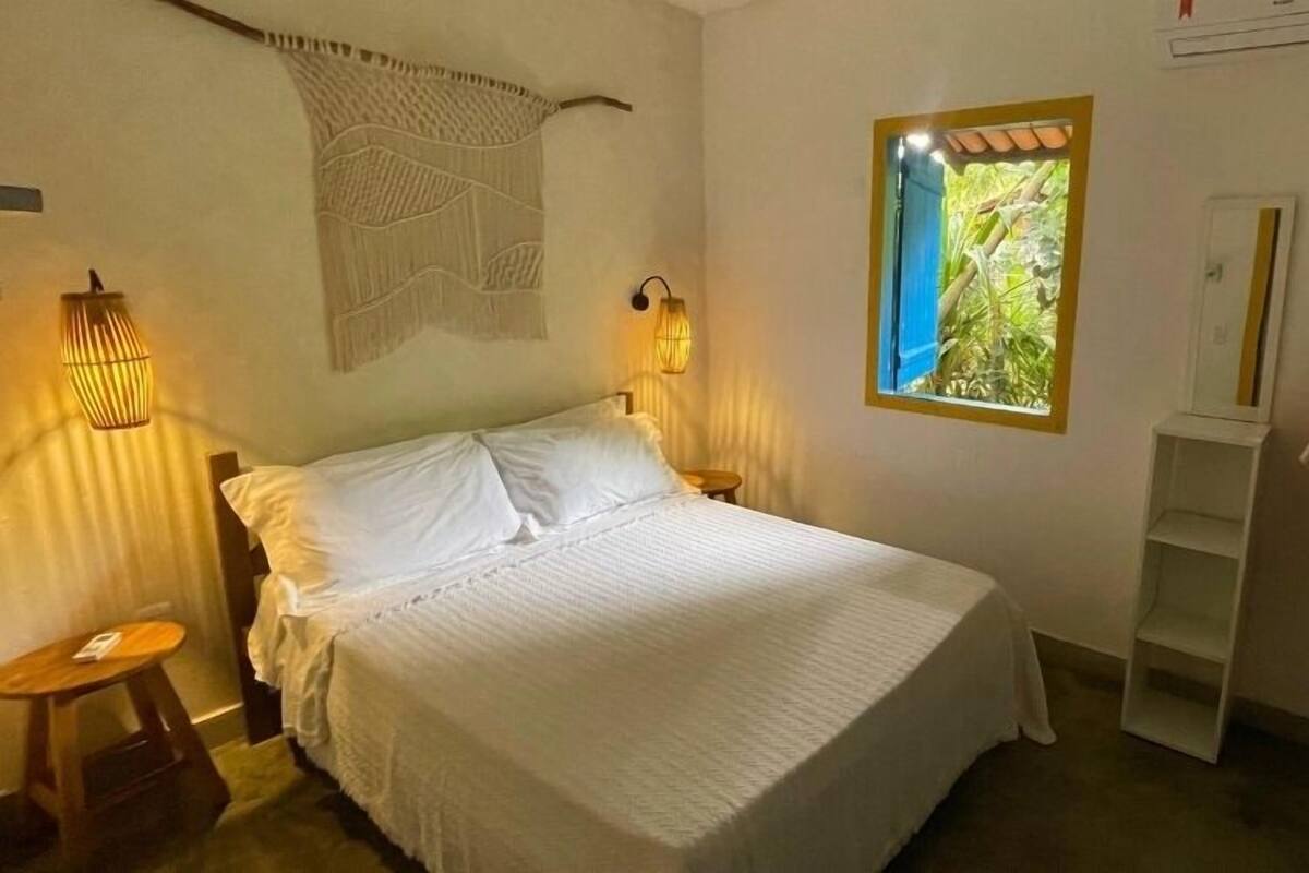 A neatly made bed with white linens, a bedside table with a lamp, a window with a view of greenery, and a clothing rack with a few items.