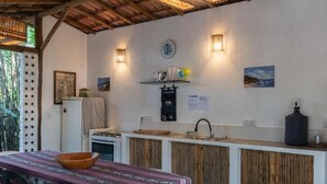 A compact kitchen with a wooden counter, a white refrigerator, and a dining area with a patterned tablecloth. - Vila Coco Dendê (Maraú)