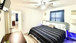 Een slaapkamer met een bed, een plafondventilator, een televisie en een raam met gordijnen.