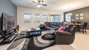 Ein modernes Wohnzimmer mit einer Ledersofa, einem Couchtisch und einem Fernseher. Es gibt große Fenster mit Jalousien, einen Deckenventilator und einen Essbereich mit Stühlen und einem Tisch.