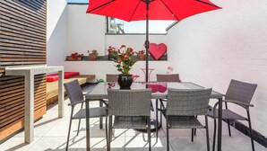 Un patio al aire libre con una mesa y sillas, un paraguas rojo, plantas en macetas y un adorno con forma de corazón en la pared.