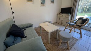 Olohuoneessa on sohva, pieni pöytä, rottinkituoli, televisiotaulutaso ja seinällä on kehystettyjä kuvia.