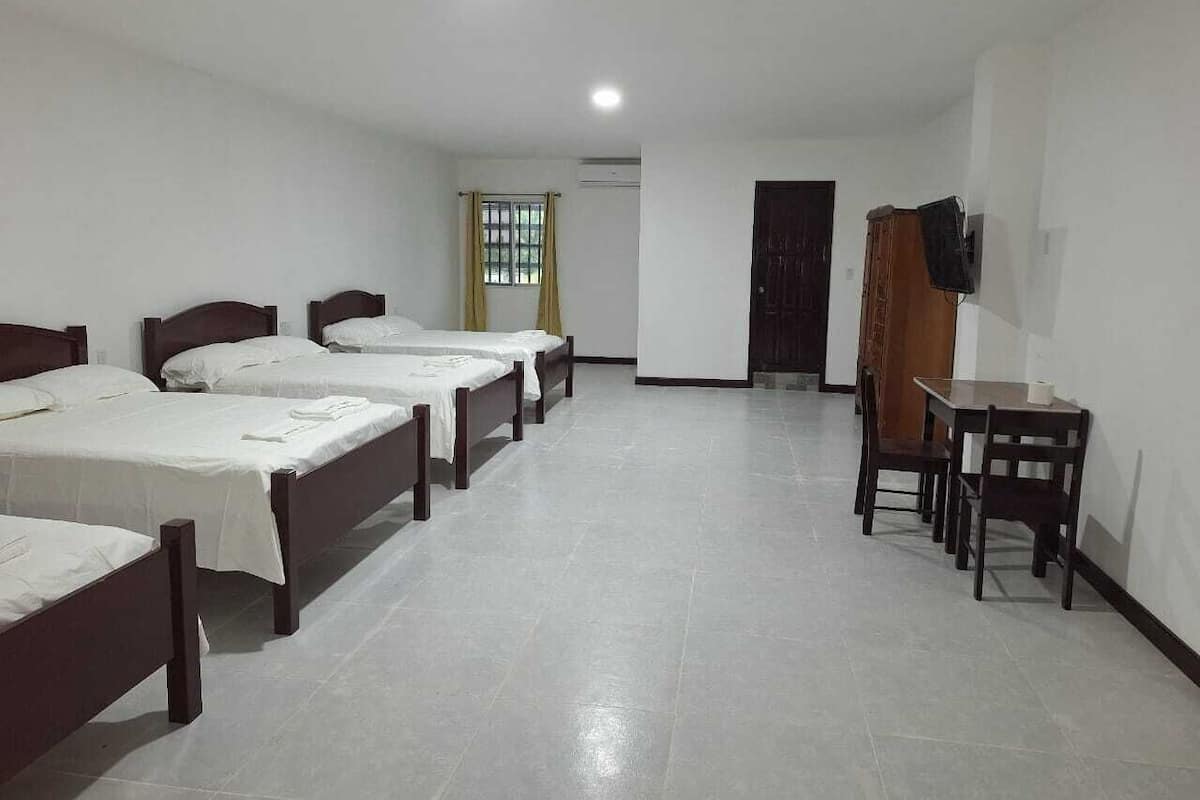 Habitación de hotel con cuatro camas individuales, una mesita con sillas, un televisor montado en la pared y una puerta que da a otra habitación.