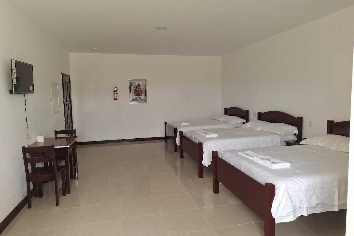 Habitación de hotel con tres camas individuales, un escritorio con sillas, un televisor y un extintor.