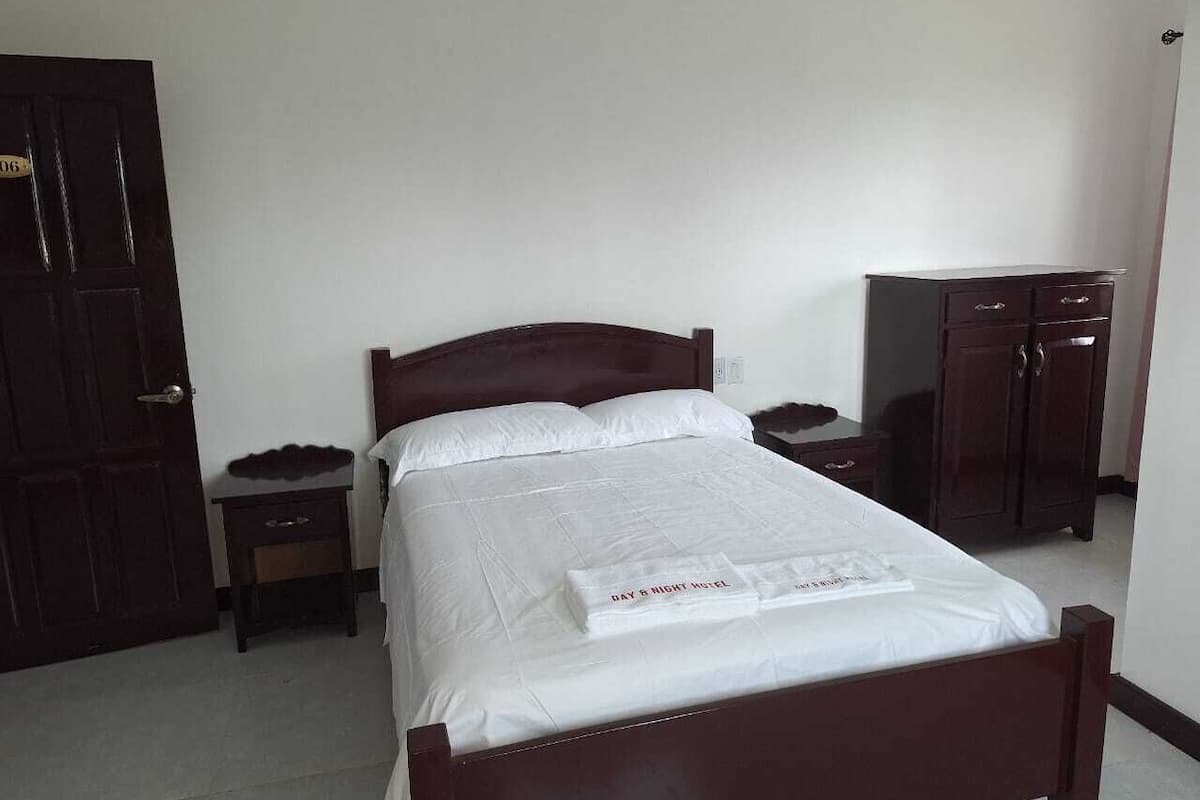 Una cama bien hecha con edredón blanco y una toalla con la leyenda "DAY & NIGHT HOTEL". Junto a la cama, una mesita de noche de madera con una lámpara, y un armario de madera oscura contra la pared.