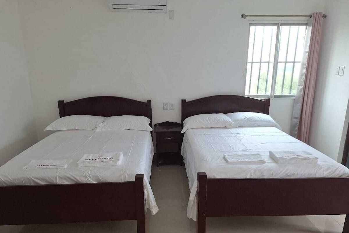 Una habitación con dos camas de madera, ropa blanca de cama, una mesita de noche pequeña y una ventana con cortinas.
