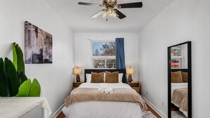 Ein Schlafzimmer mit einem Bett, einem Deckenventilator, einem Fenster mit Vorhängen, einem Nachttisch mit Lampe und einem großen Spiegel.
