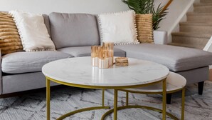 Ein modernes Wohnzimmer mit einem grauen Sofa, einem runden Couchtisch mit goldenen Beinen und einer Topfpflanze.