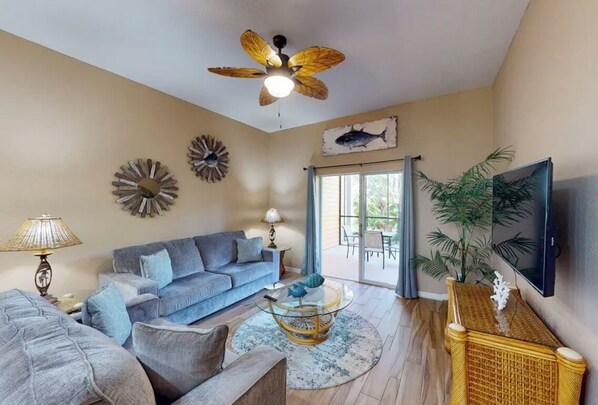 A living room with a blue sofa, a round coffee table, a ceiling fan, and a television mounted on the wall.