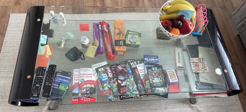 Uma mesa com tampo de vidro e diversos objetos, incluindo controles remotos, livros e uma cesta de frutas.