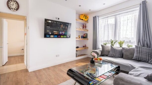 A modern living room with a flat-screen TV mounted on the wall, a sofa with cushions, a coffee table with books, and a window with curtains. - Fresh Apartment & Parking (London)