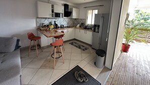 A modern kitchen with white cabinets, a central island with stools, a refrigerator, and a window with a view of greenery. - Residence La Terrasse (POINTE NOIRE)