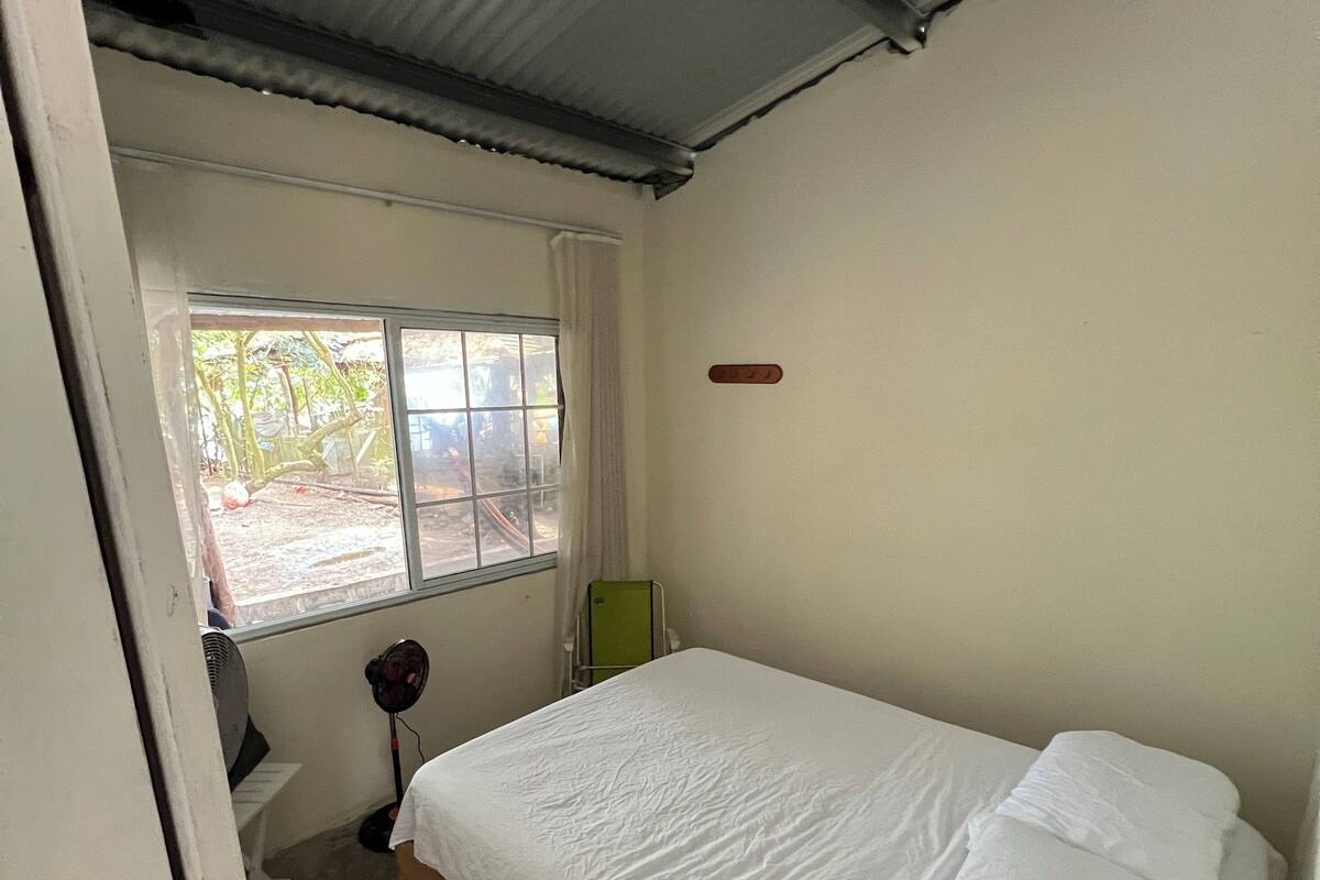 A small, single-bed room with a window, a fan, and a green chair.