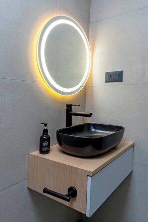 Un baño moderno con un lavamanos tipo cuenco negro, un mueble de tocador de madera, un dispensador de jabón negro y un espejo circular con luz.