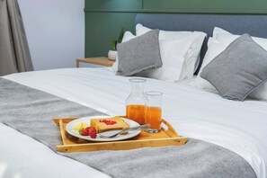 Una cama bien hecha con un edredón gris, una bandeja de madera con cosas para el desayuno y una mesita de noche con una planta.