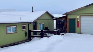 Propiedad cubierta de nieve con terraza de madera, garaje y un pequeño edificio con una puerta marcada '821'.
