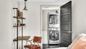 Een compacte slaapkamer met een bed, een klein houten tafeltje, een bruine leren fauteuil, een ingebouwde wasmachine en droger, en een donkere deur.