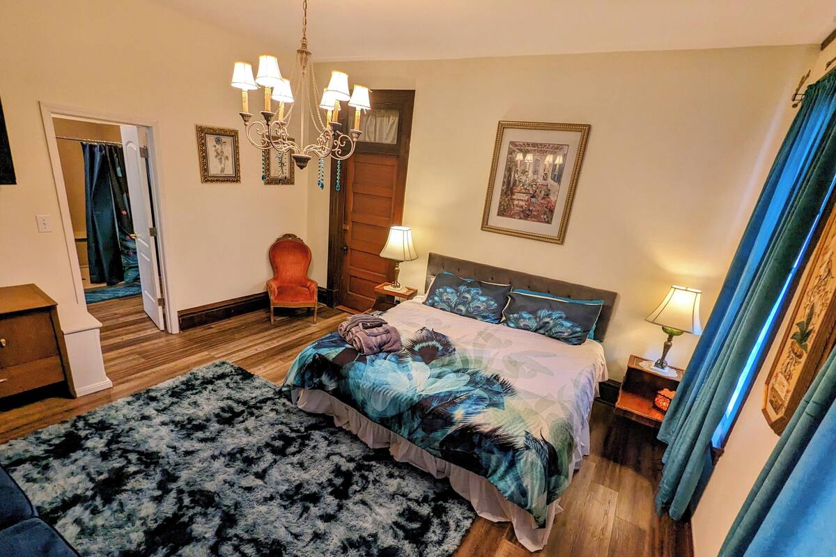 A bedroom with a bed, a chair, a chandelier, a rug, and a door.