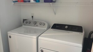 A laundry room with two white Whirlpool washers and dryers, a shelf above containing cleaning supplies, and a power outlet on the wall.