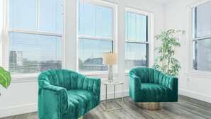 A room with two teal armchairs, a small side table, and a floor lamp. There are large windows with blinds, and a potted plant is visible.