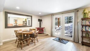 A dining area with a wooden table and chairs, a red fireplace, and a large window with a view of snow-covered trees.