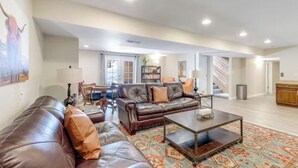 A spacious living room with a brown leather sofa, a coffee table, and a patterned rug. There is a dining area with a table and chairs in the background.