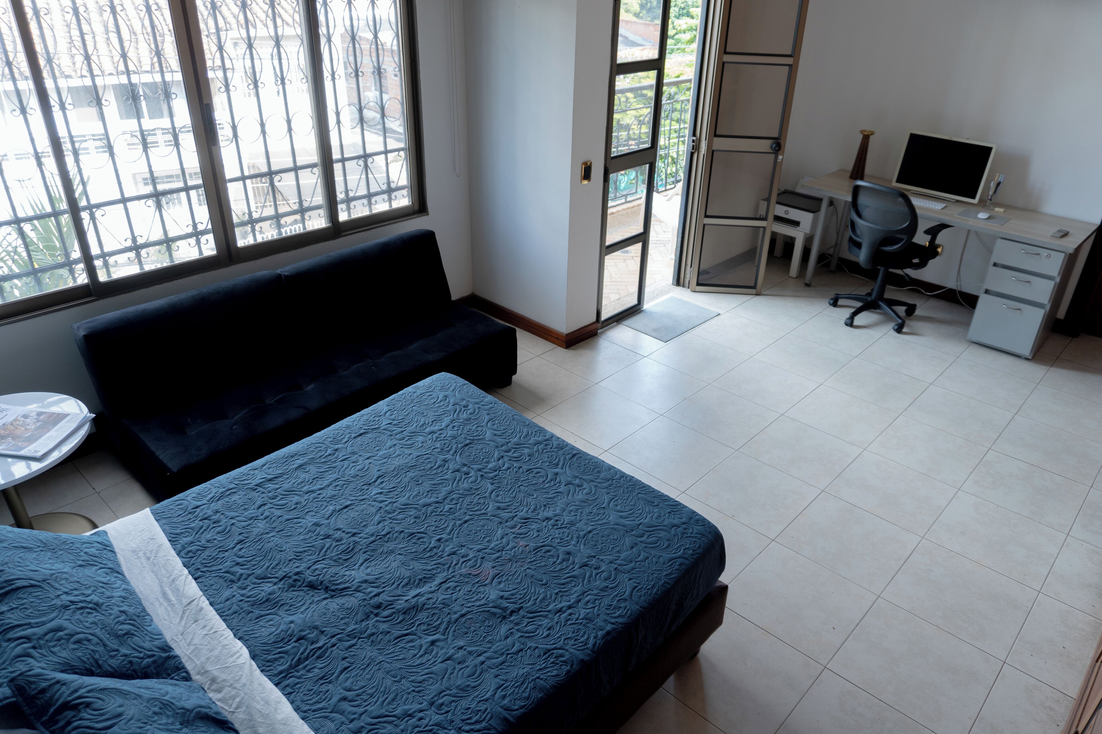 Een kamer met een bed, een bureau en een computer. Er is een raam met metalen tralies en een deur naar een buitenruimte.