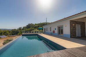 A swimming pool with a wooden deck, a white building with a stone facade, and solar panels in the background.