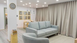 Un salon moderne avec un canapé bleu clair, une table d’appoint en bois sur laquelle sont posés des livres, une horloge ronde et, au fond, un coin repas.