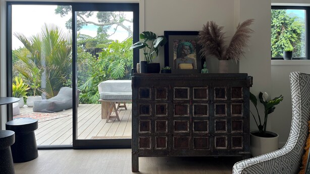 Un salon moderne doté d’une porte-fenêtre coulissante donnant sur une terrasse en bois, d’un fauteuil à motifs et d’un meuble décoratif agrémenté de plantes.