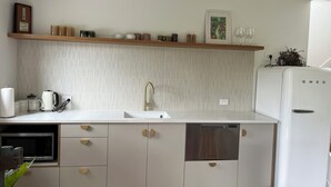 Une cuisine moderne avec des meubles blancs, un four à micro-ondes en acier inoxydable, un réfrigérateur blanc et une étagère en bois au-dessus de l’évier.