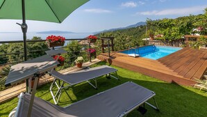 A poolside area with a view of the sea and mountains, a wooden deck, and lounge chairs.