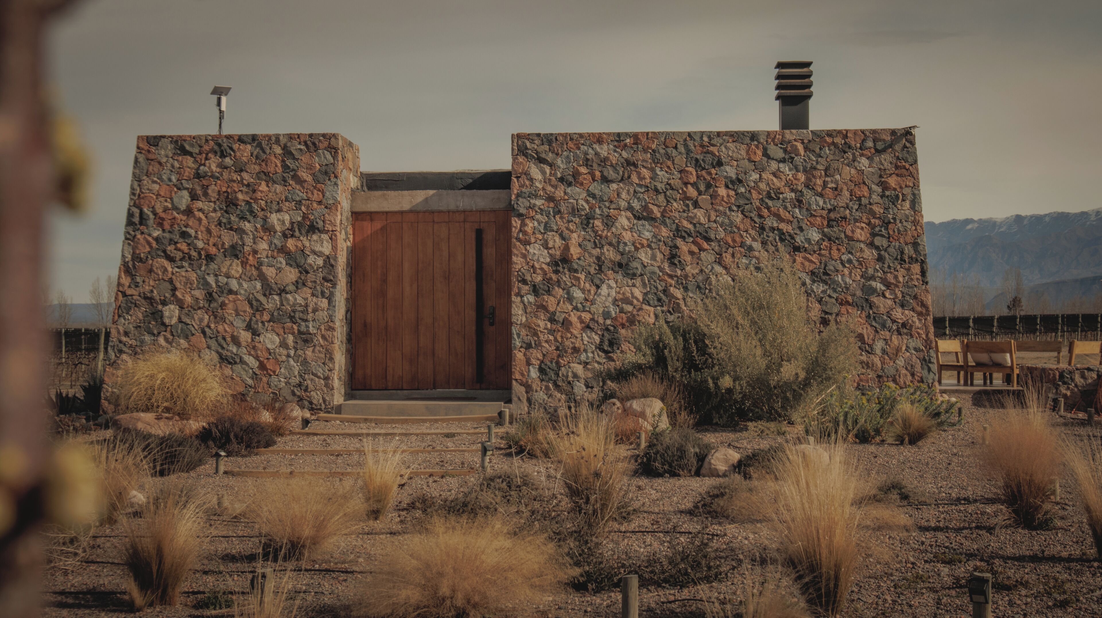 A rustic building with a wooden door and stone walls, surrounded by dry vegetation and a mountainous backdrop.