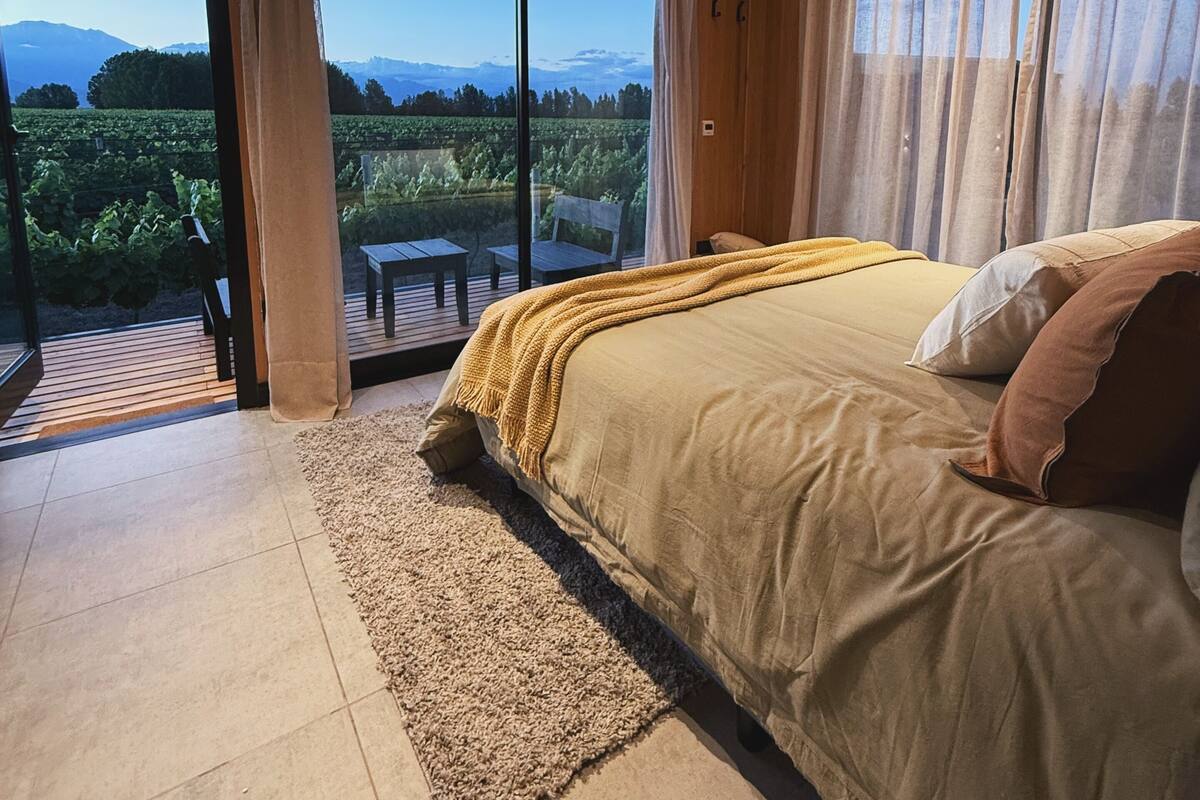 A bedroom with a large bed, a wooden ceiling, and a view of a vineyard through large windows.