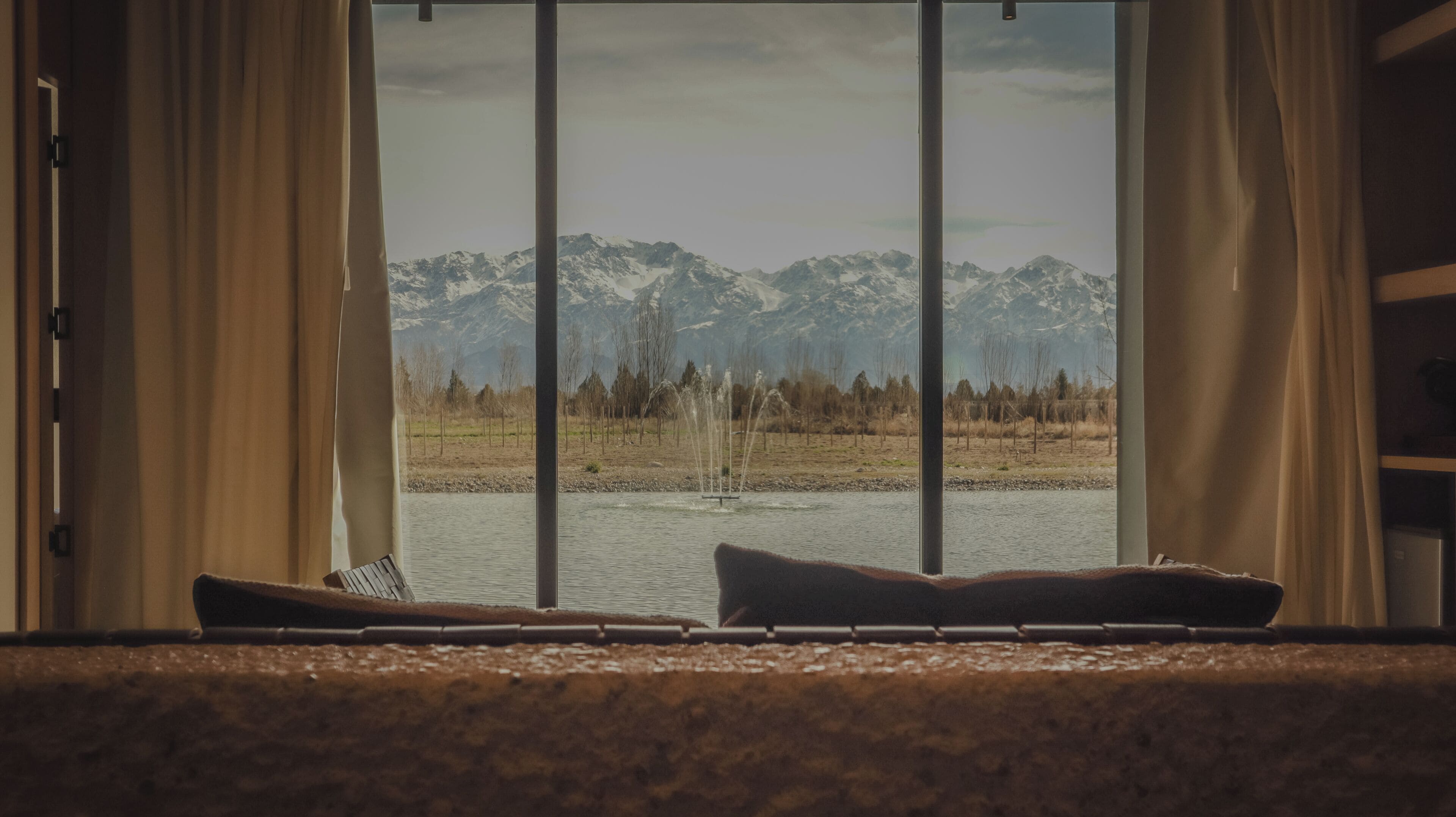 Ein Zimmer mit großem Fenster und Blick auf eine gebirgige Landschaft, ein Bett mit Kissen sowie ein kleiner Tisch.
