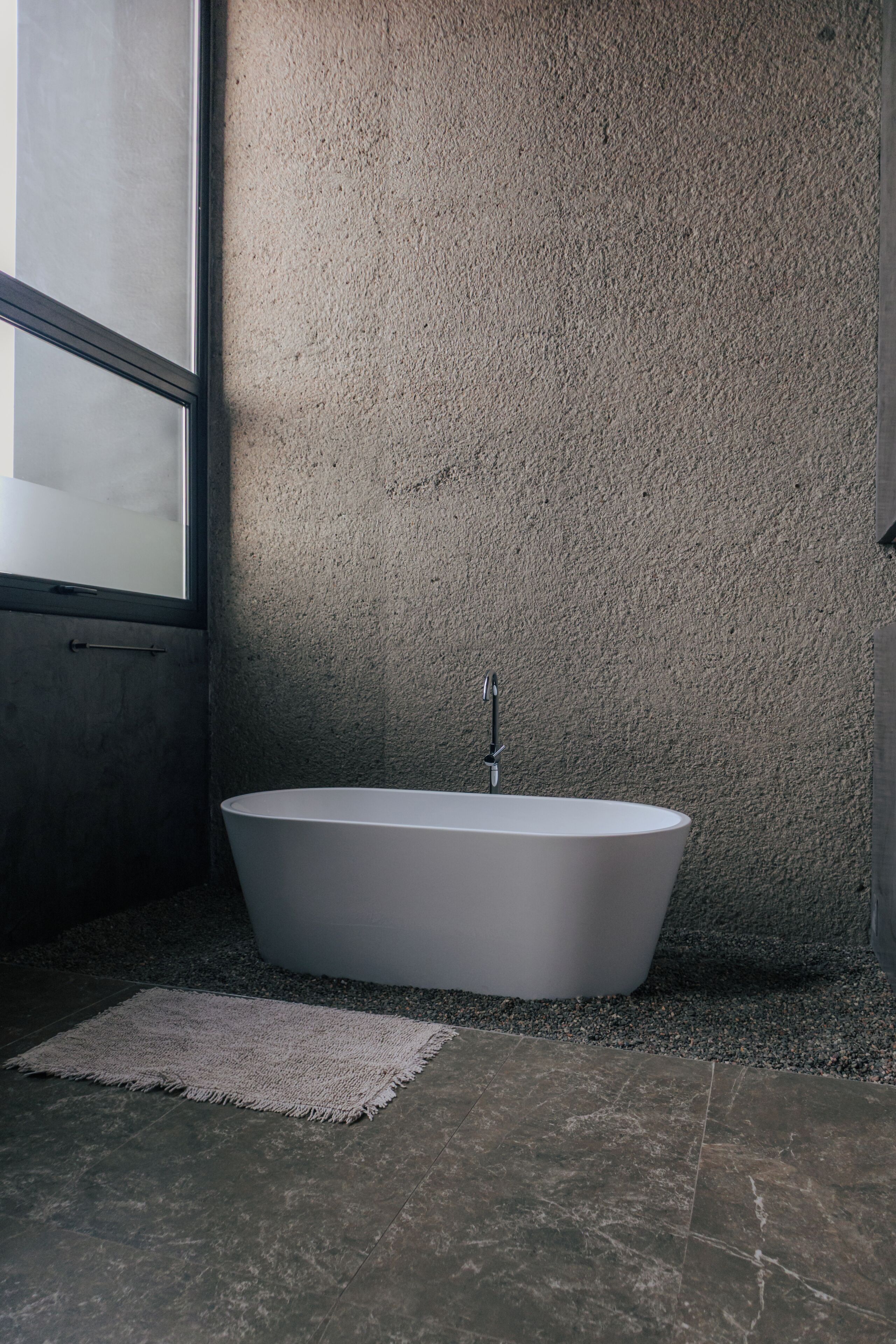 A modern bathroom with a freestanding bathtub, a textured wall, and a large window.