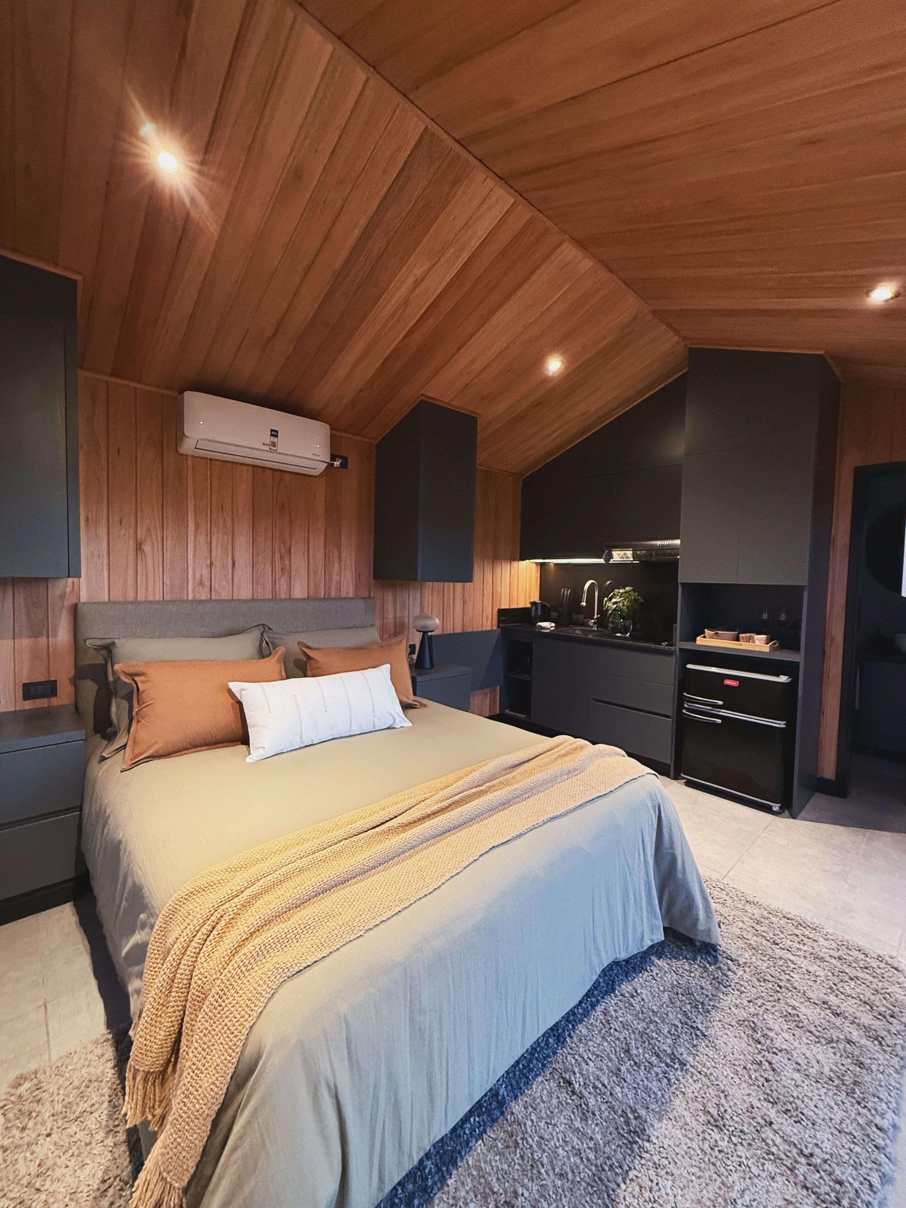 A modern bedroom with a wooden ceiling, a large bed with a beige and blue bedspread, a small bedside table, and a built-in TV cabinet.