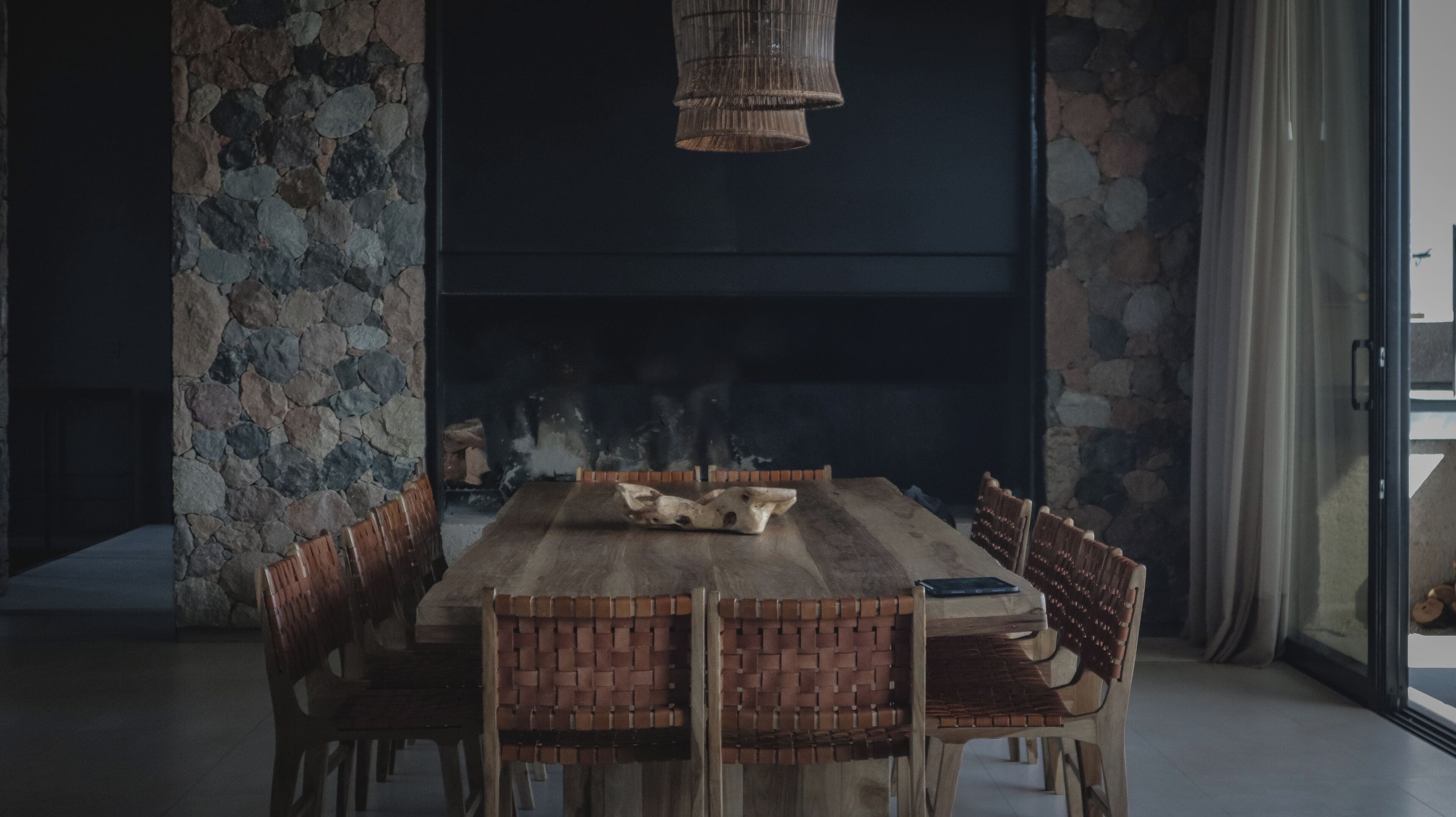 A dining area with a stone wall, a wooden table, and woven light fixture.