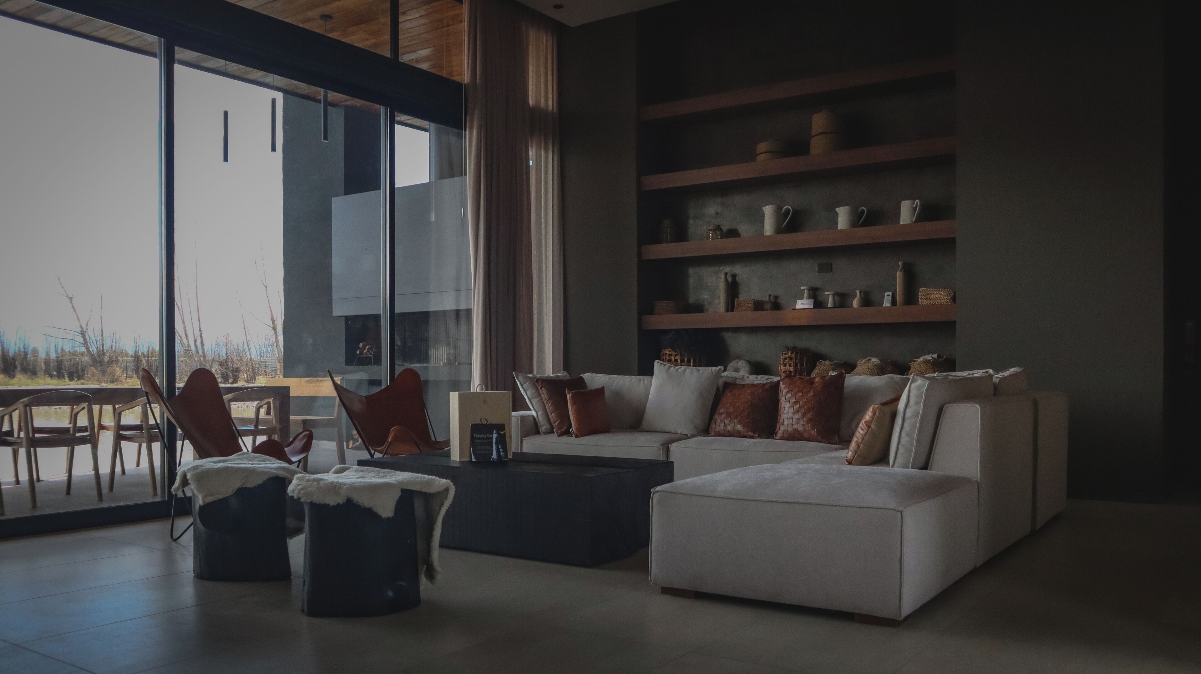 A modern living room with a sofa, chairs, and a coffee table. There are shelves with decorative items and a view of the outdoors through large windows.