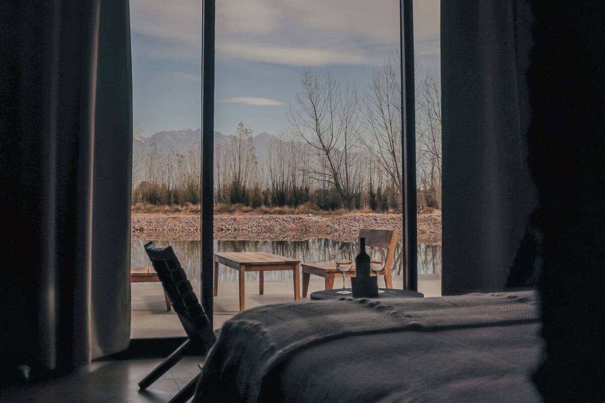A bedroom with a large window, a bed, a chair, and a table with a bottle.