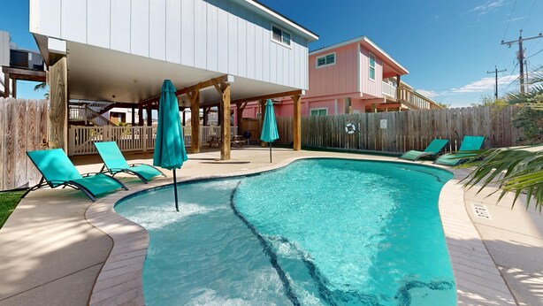 Una piscina con sillas de descanso y sombrillas, una terraza de madera y una casa con porche cubierto.