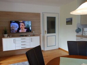 Un soggiorno moderno con TV a schermo piatto, un mobiletto in legno, un tavolo da pranzo con sedie e un angolo cottura con macchina da caffè.