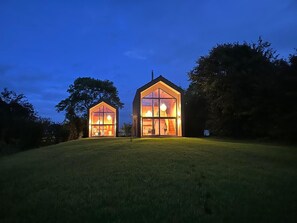 Two modern, glass-fronted houses with visible interior lighting, set on a grassy hill with trees in the background.