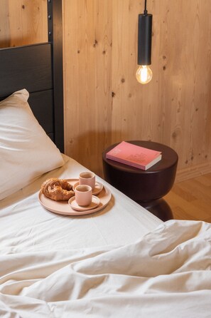 Cama bem arrumada com uma bandeja de croissants e xícaras de café, um livro em um banquinho e uma lâmpada pendente.