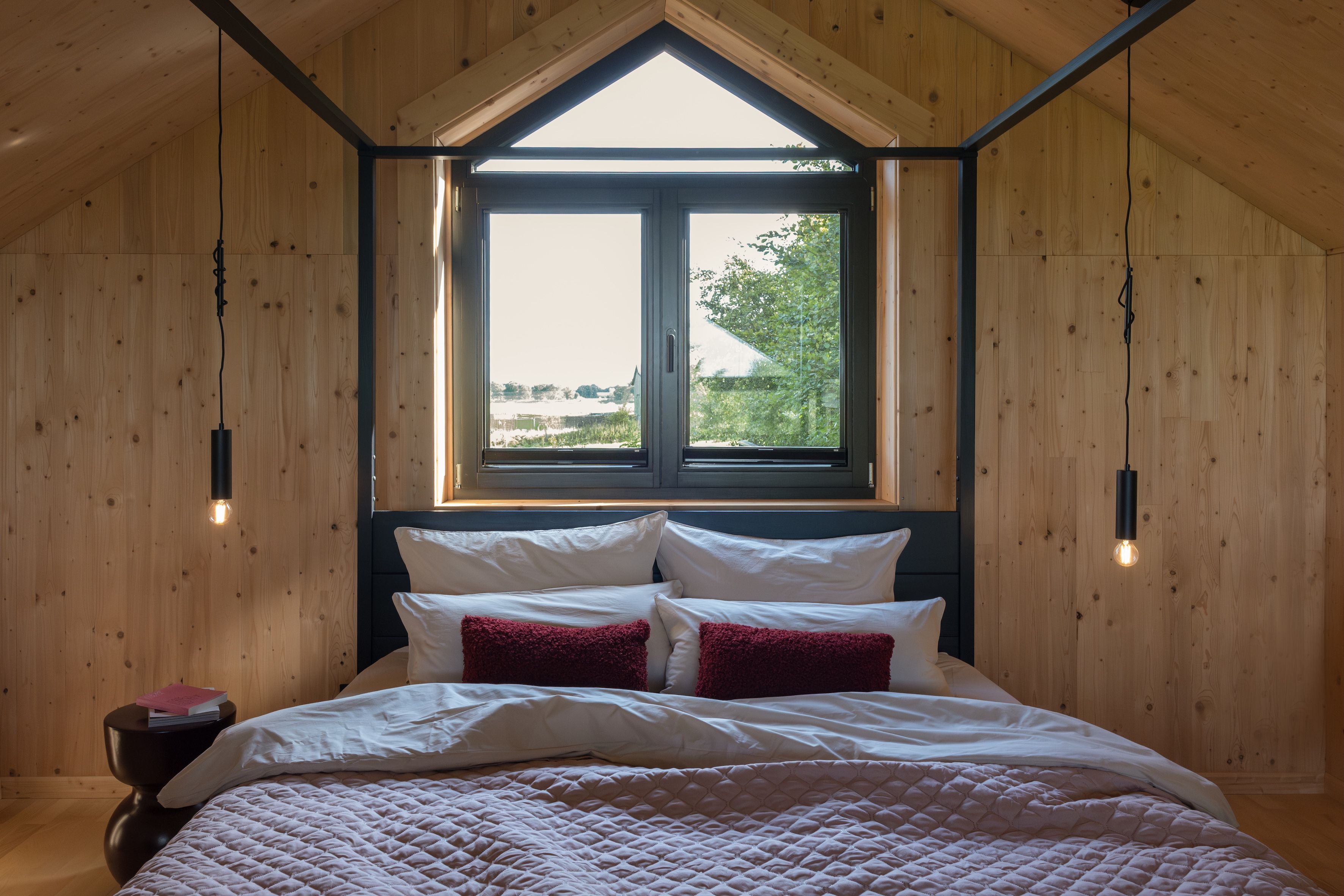 Ein Schlafzimmer mit Holzboden, einem großen Fenster und einem Bett mit weißen und roten Kissen.