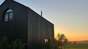 A large, dark wooden barn with a single window and chimney, surrounded by a grassy field and a tree.