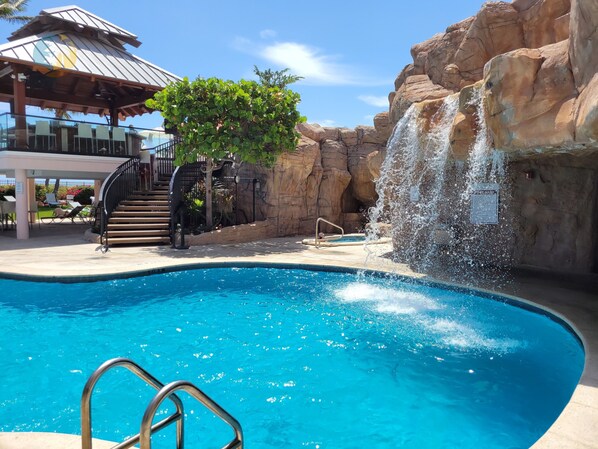 A swimming pool with a waterfall feature, surrounded by rock formations and a covered seating area with lounge chairs.