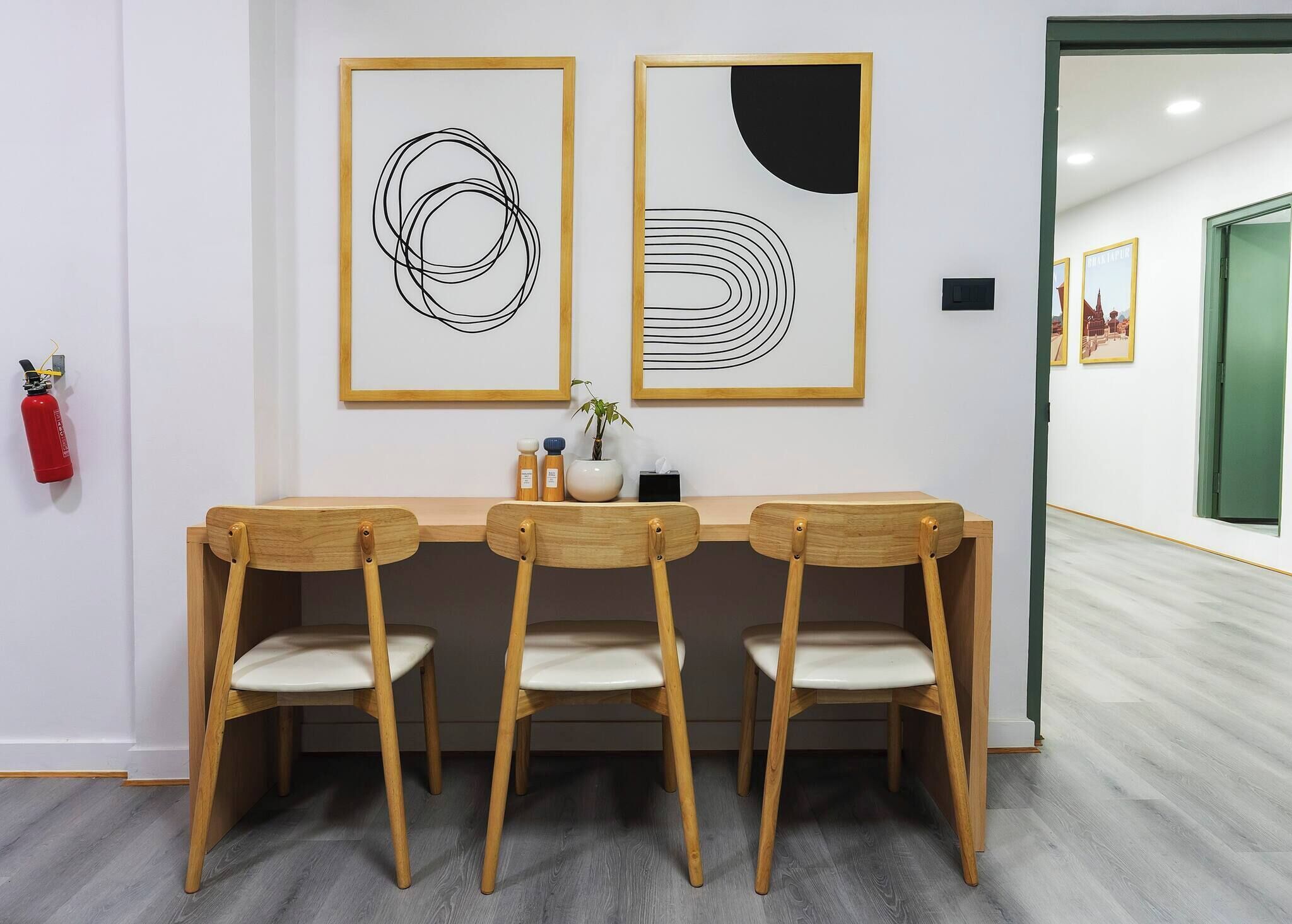 A dining area with a wooden table and chairs, two abstract paintings on the wall, a fire extinguisher, and a green door in the background.