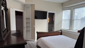 Une chambre à coucher comprenant un lit, une commode, un téléviseur fixé au mur et une fenêtre avec des stores.