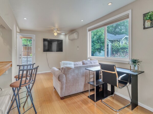 A compact living space with a sofa, a small table, a chair, a TV mounted on the wall, and a window with a view of greenery.